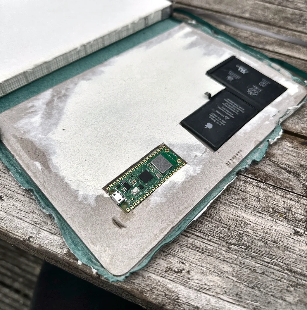 An early build stage outdoors on a weathered wooden bench: the notebook's board covers split open and hollowed, with filler smoothed into the recess, and a Raspberry Pi Pico and two flat battery cells laid in place to check they fit.