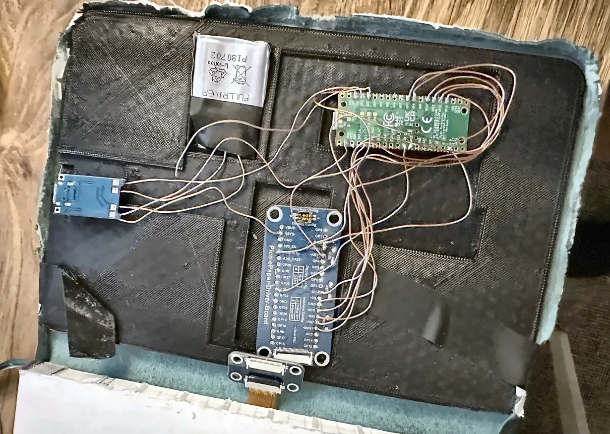 The inside back cover of a hardback notebook, hollowed out and lined with black moulded trays. Set into it are a lithium-polymer pouch cell, a small blue charging module, a Raspberry Pi Pico and a Pico e-paper driver board, wired together with fine enamelled copper wire, with a ribbon cable running out to the display.