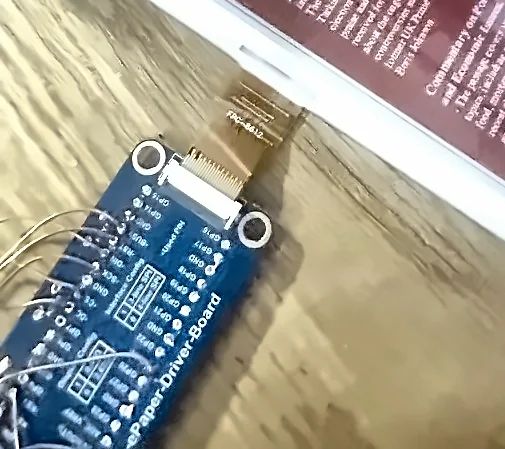 Close view of the blue Pico e-paper driver board with its GPIO labels visible, a flat ribbon cable running from its connector into the edge of the e-paper panel behind it.