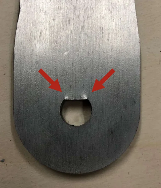 Close-up of a machined metal crank arm with a rounded end and a pin slot, with two red arrows pointing at cracks propagating from either side of the slot.