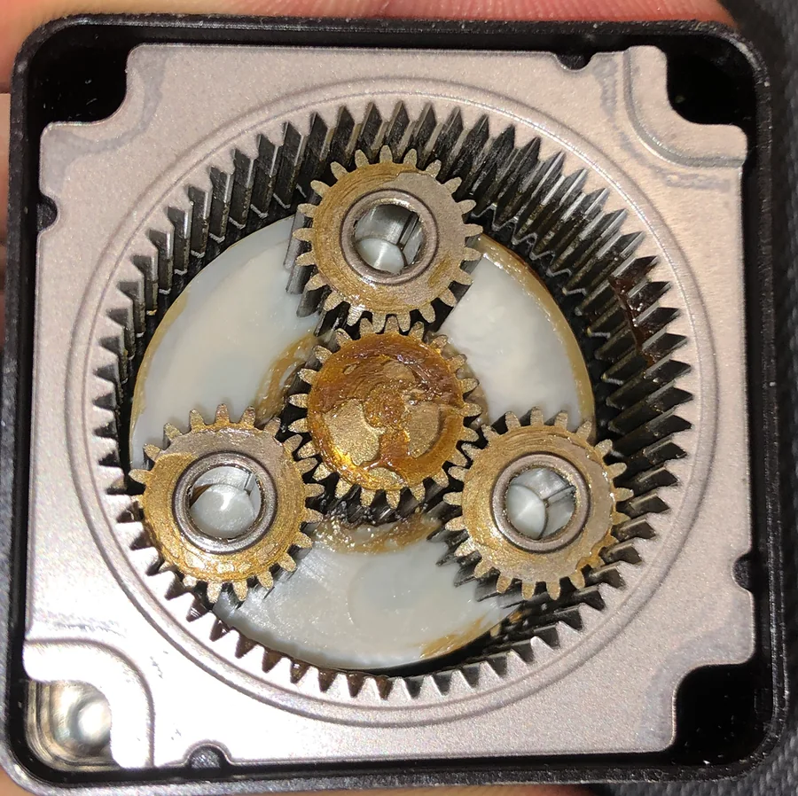 An opened planetary gearbox photographed from above, showing a large internal ring gear, three planet gears and a central sun gear, with grease spread across the faces and visible wear on the tooth flanks.