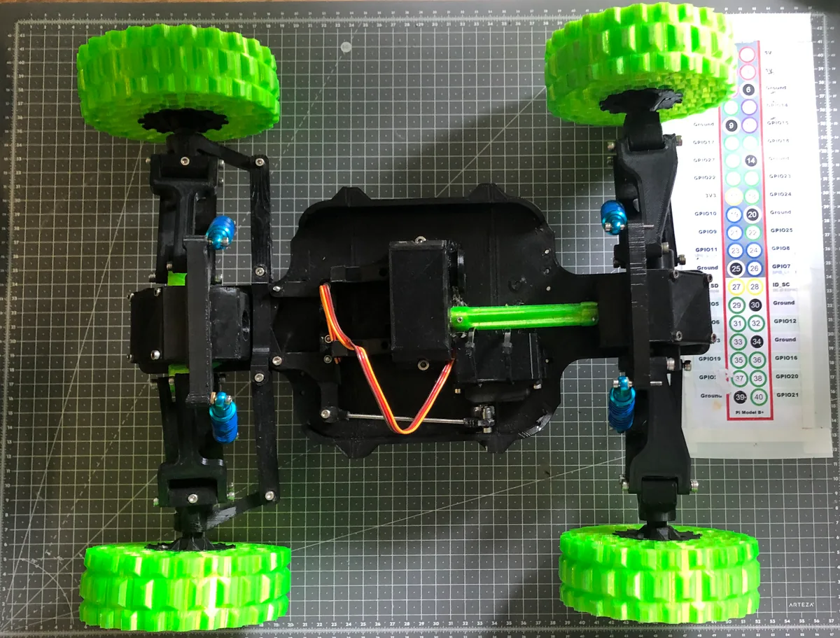The rover chassis on a cutting mat during assembly, with a Raspberry Pi GPIO pinout reference card beside it and servo wiring routed through the frame.