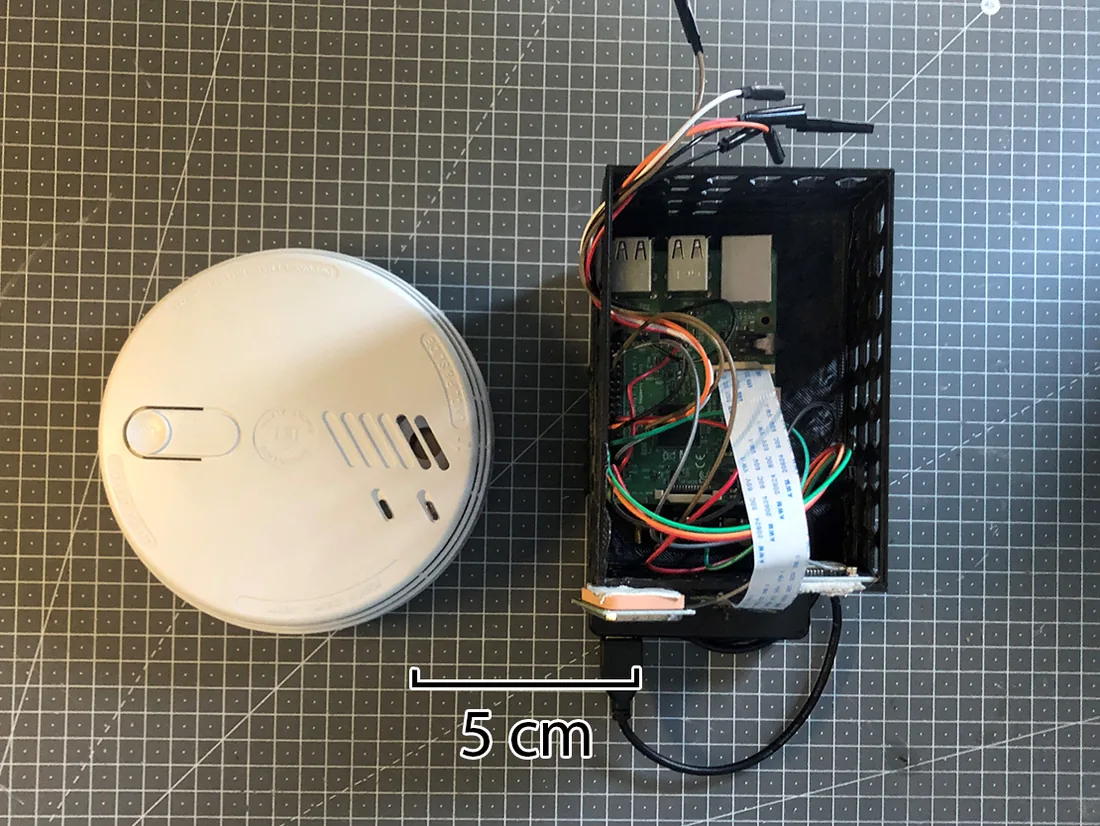 The sensor unit, a Raspberry Pi and Geiger board in a printed vented case with ribbon and jumper wiring, photographed next to a domestic smoke detector with a five centimetre scale bar.