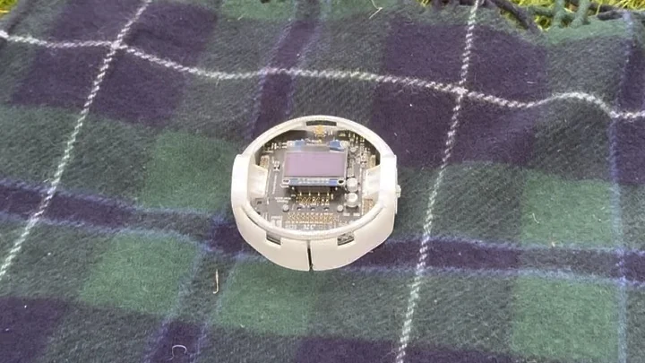 A Pololu 3Pi+ robot, a small white two-wheeled disc with an exposed circuit board and an OLED display, sitting on a tartan blanket