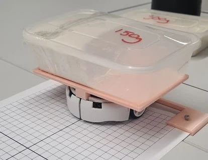 A sealed food container labelled 150 g strapped to a printed platform over one side of the robot, offsetting the load to that wheel.