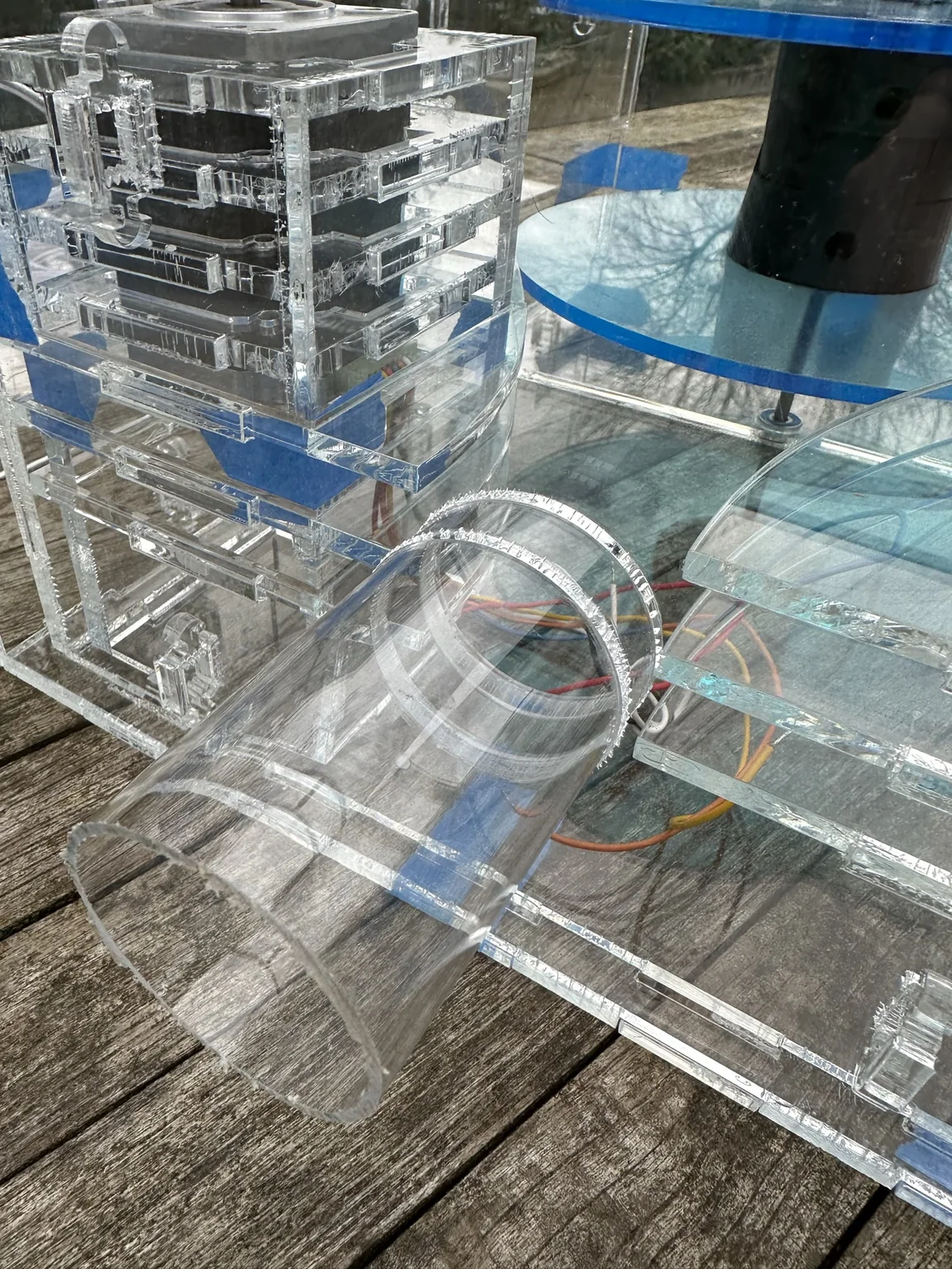 The second launcher in clear acrylic on a wooden deck: the stepper motor in its laser-cut mount, the blue spool discs behind it, and the clear exit tube at the front.