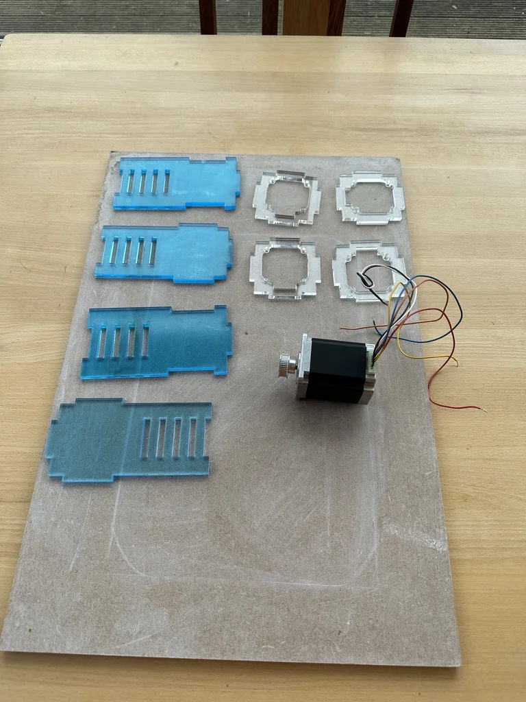 Laser-cut acrylic parts laid out on an MDF board, four blue side panels and four clear motor-mount frames, next to a stepper motor with its wiring loose.